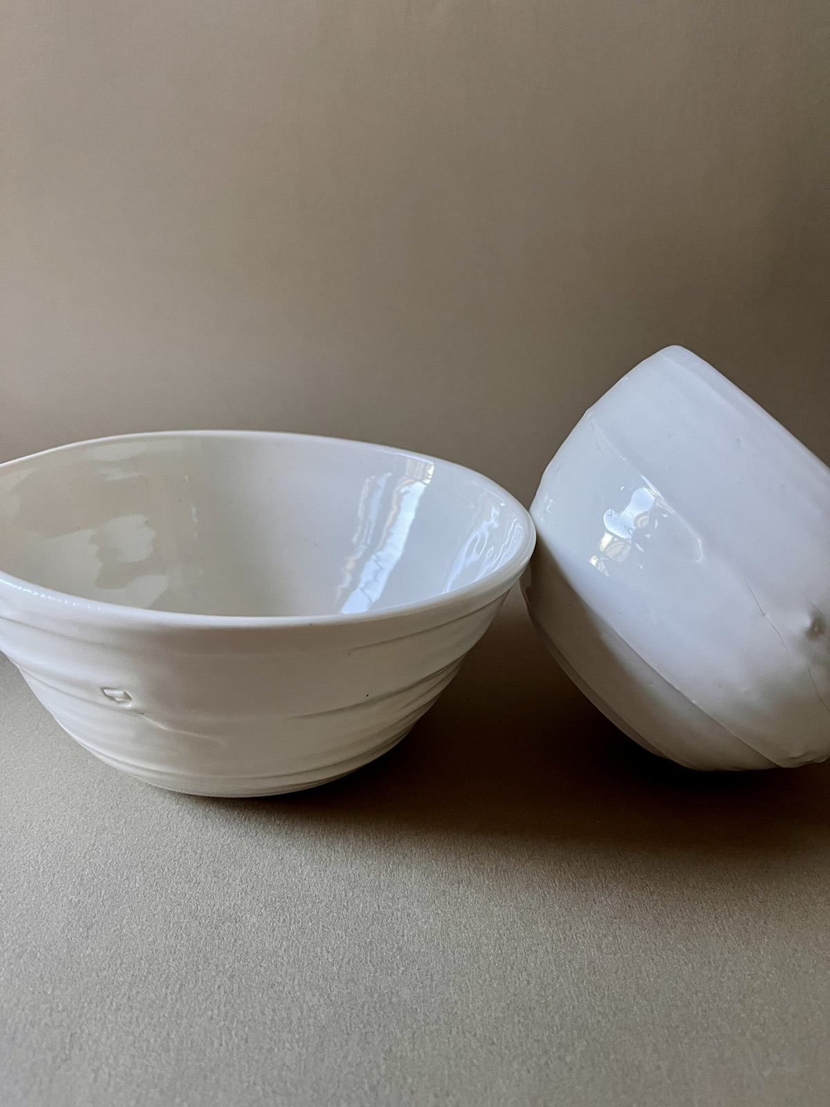 Deux bols Purete en porcelaine blanche brillante, l'un appuyé contre l'autre, sur fond beige