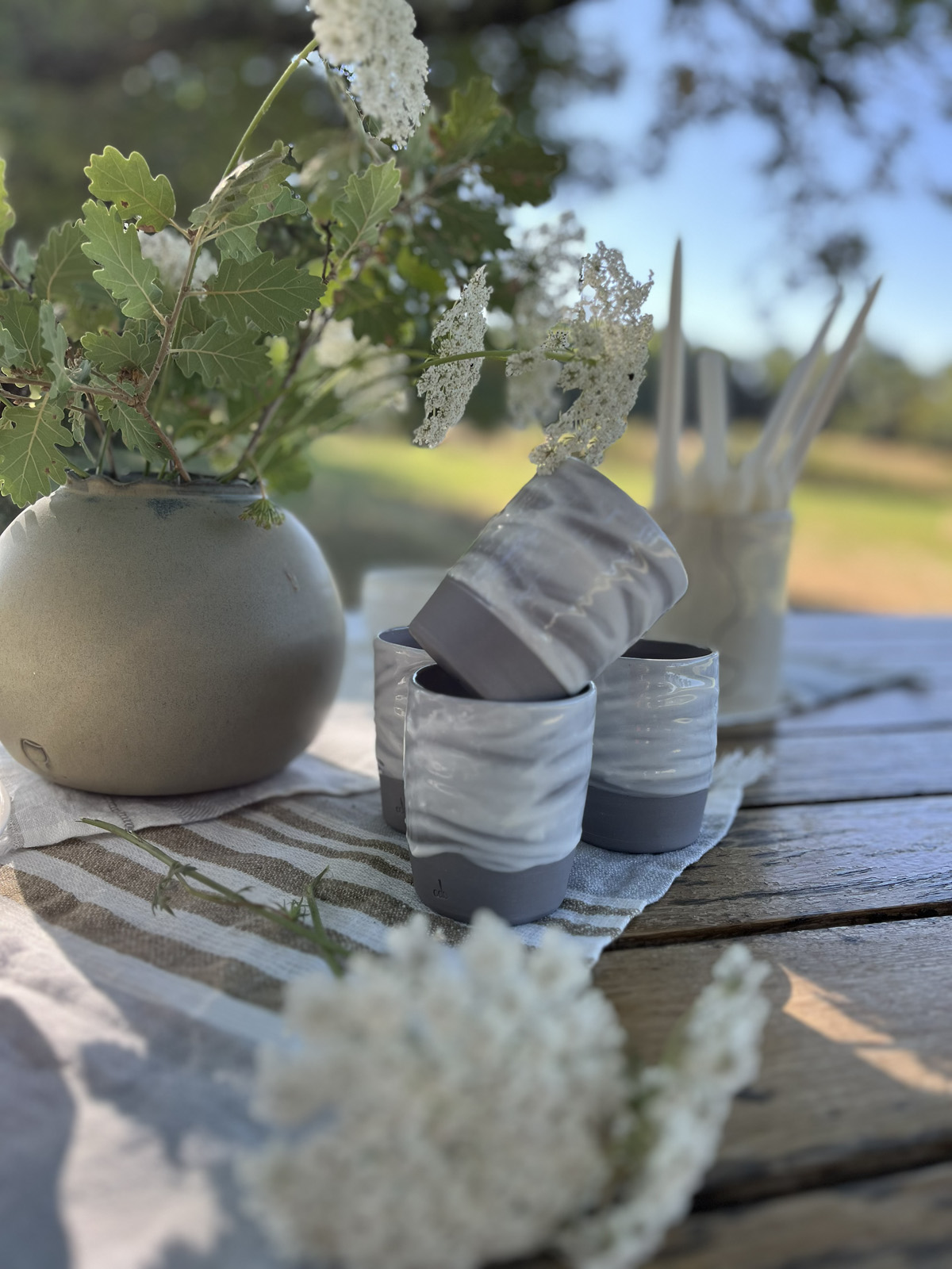 Gobelets en grès marbré gris et blanc et vase rond en grès garni de fleurs sauvages, sur une table en bois
