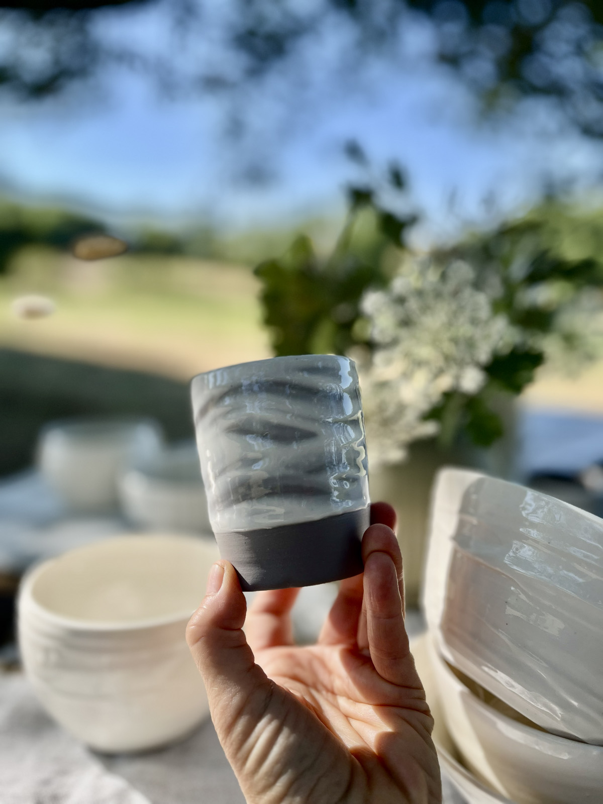 Gobelet en grès marbré gris et blanc à base noire tenu en main, bols blancs et fleurs en fond