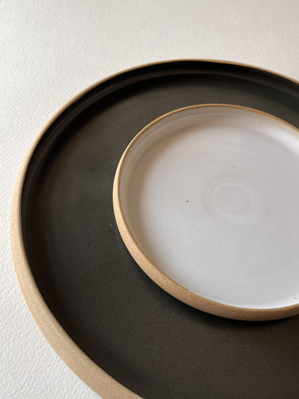 Petite assiette blanche brillante posée sur une grande assiette noire mate, bords en grès brut