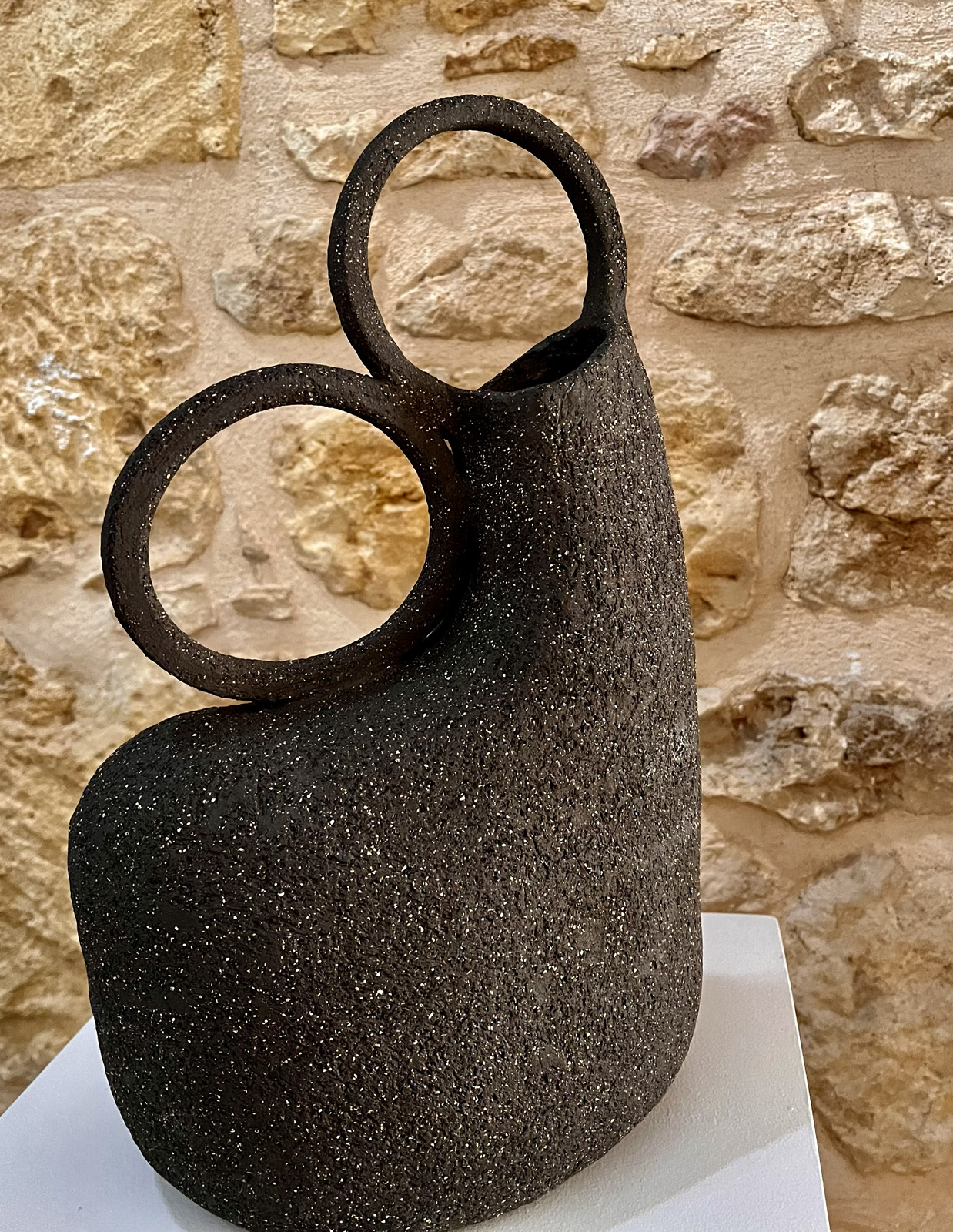 Vase sculptural en grès noir chamotté à deux anneaux vu sous un autre angle, mur de pierre en fond
