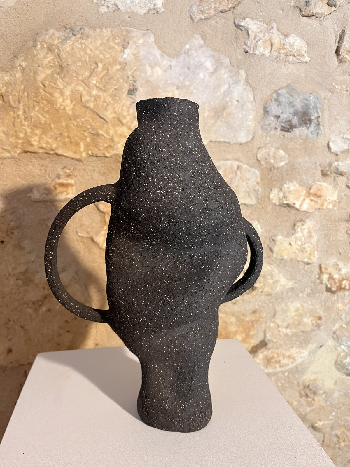 Vase sculptural en grès noir chamotté à deux anses, forme torsadée, sur socle devant un mur de pierre