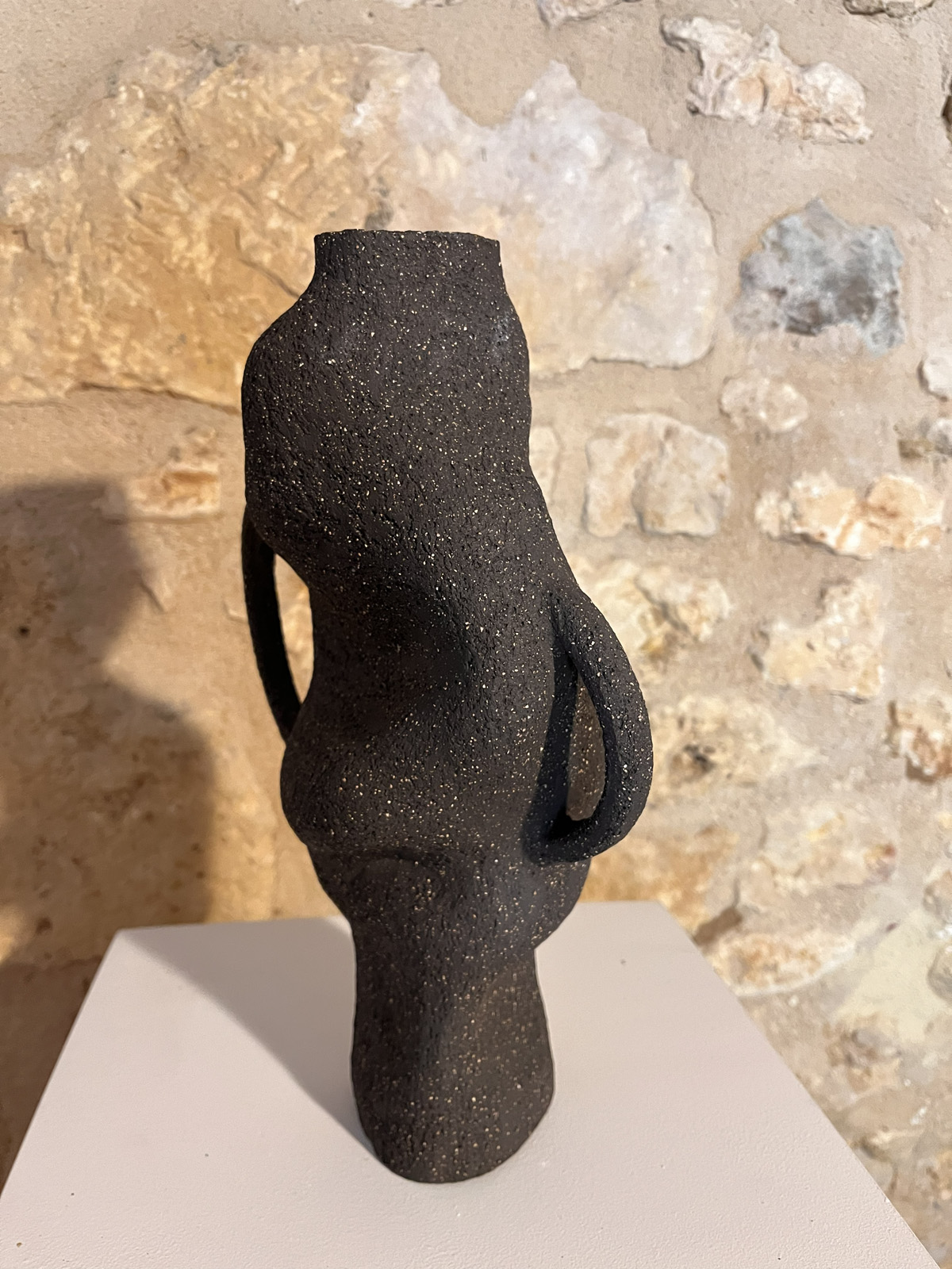 Vase sculptural en grès noir chamotté à anses, forme torsadée, vu de trois quarts devant un mur de pierre