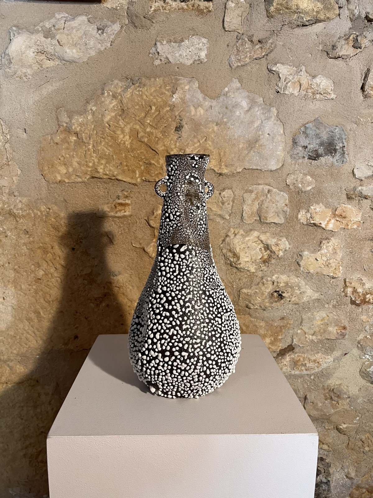 Vase Winter entièrement couvert d'émail blanc rétracté sur grès noir, sur socle devant un mur de pierre
