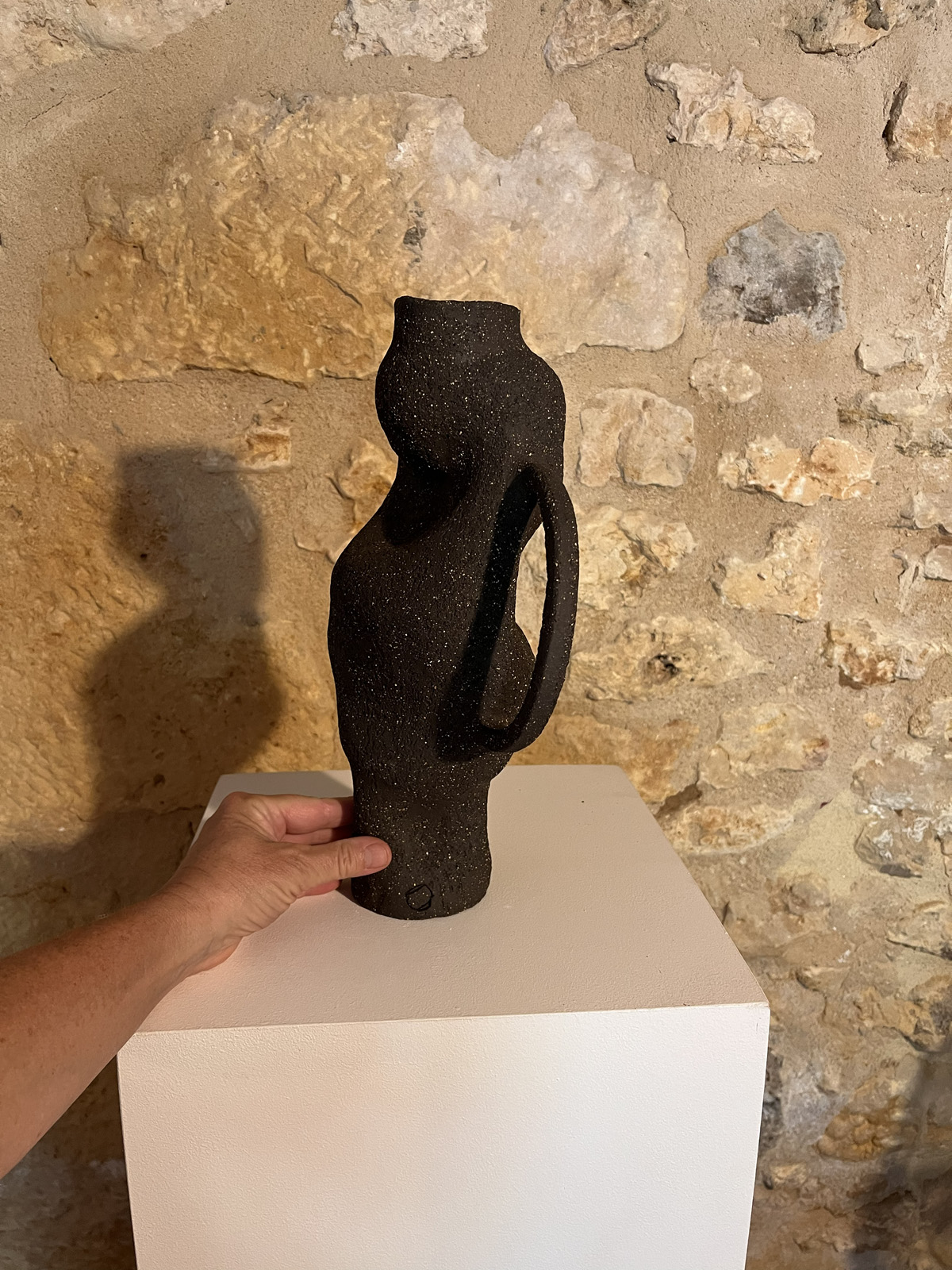 Main tenant un vase sculptural en grès noir chamotté à anse et forme torsadée, devant un mur de pierre