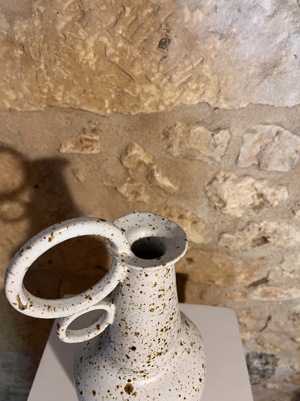 Gros plan sur le col fin et les anneaux d'un vase en grès blanc moucheté de brun, mur de pierre