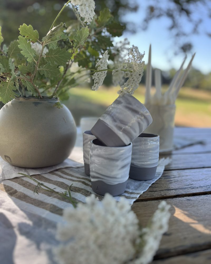 Gobelets en grès marbré gris et blanc et vase rond en grès garni de fleurs sauvages, sur une table en bois
