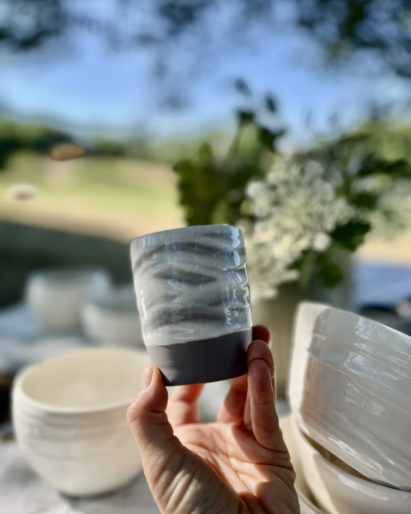 Gobelet en grès marbré gris et blanc à base noire tenu en main, bols blancs et fleurs en fond