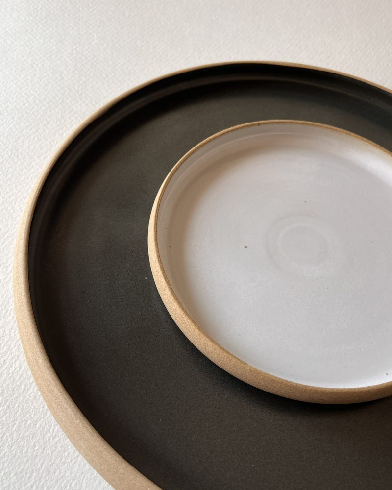 Petite assiette blanche brillante posée sur une grande assiette noire mate, bords en grès brut
