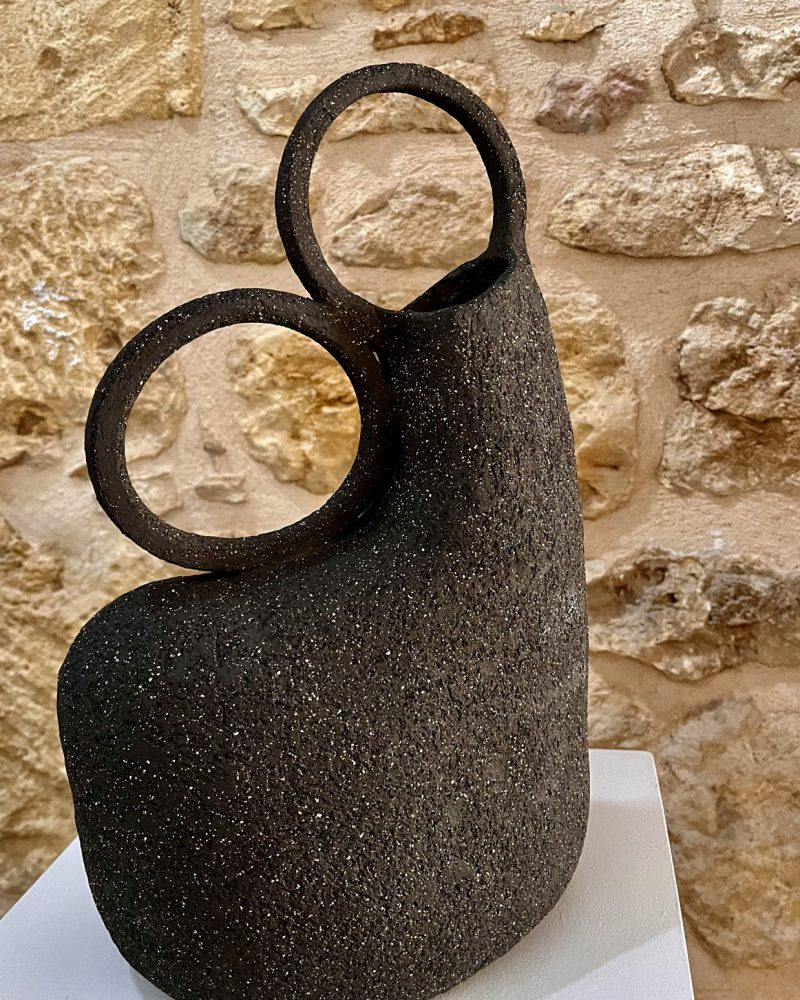Vase sculptural en grès noir chamotté à deux anneaux vu sous un autre angle, mur de pierre en fond