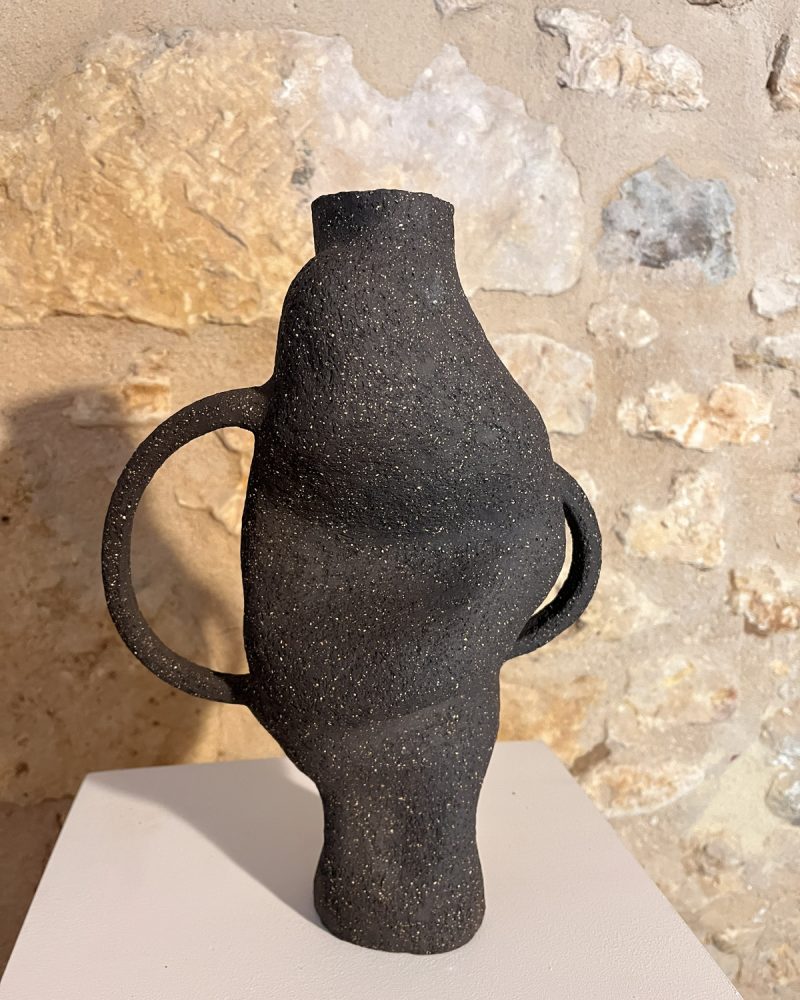 Vase sculptural en grès noir chamotté à deux anses, forme torsadée, sur socle devant un mur de pierre