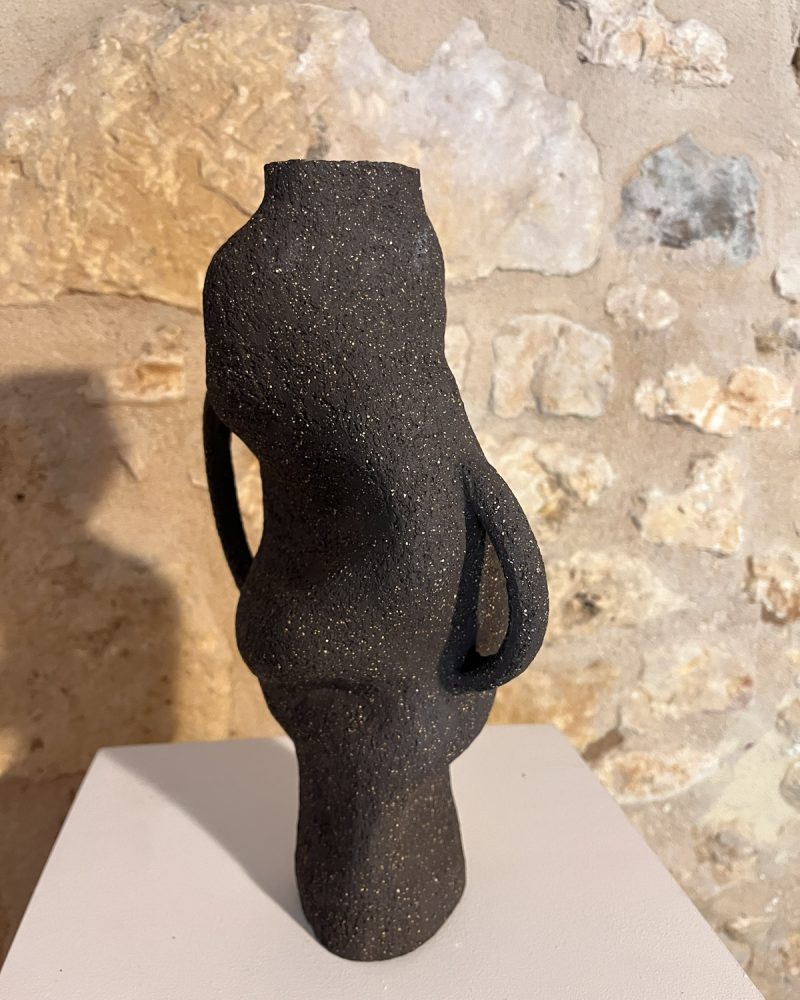 Vase sculptural en grès noir chamotté à anses, forme torsadée, vu de trois quarts devant un mur de pierre