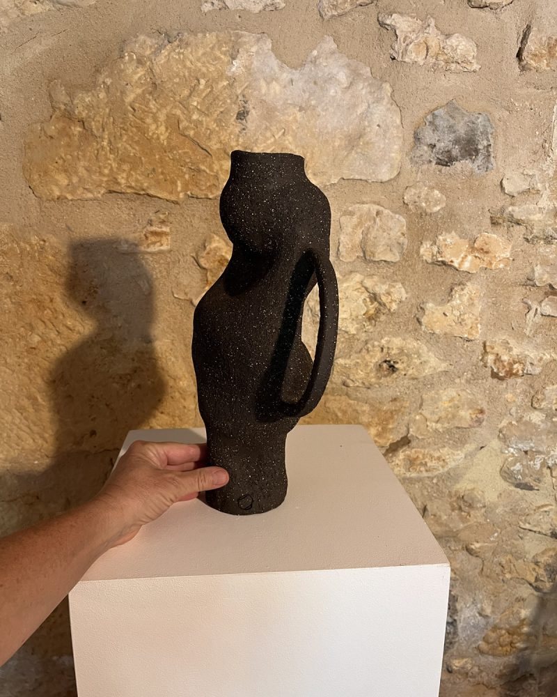 Main tenant un vase sculptural en grès noir chamotté à anse et forme torsadée, devant un mur de pierre