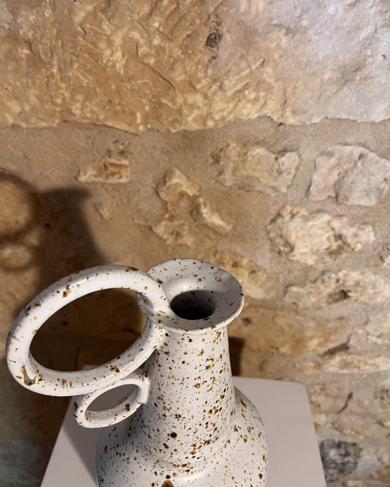 Gros plan sur le col fin et les anneaux d'un vase en grès blanc moucheté de brun, mur de pierre